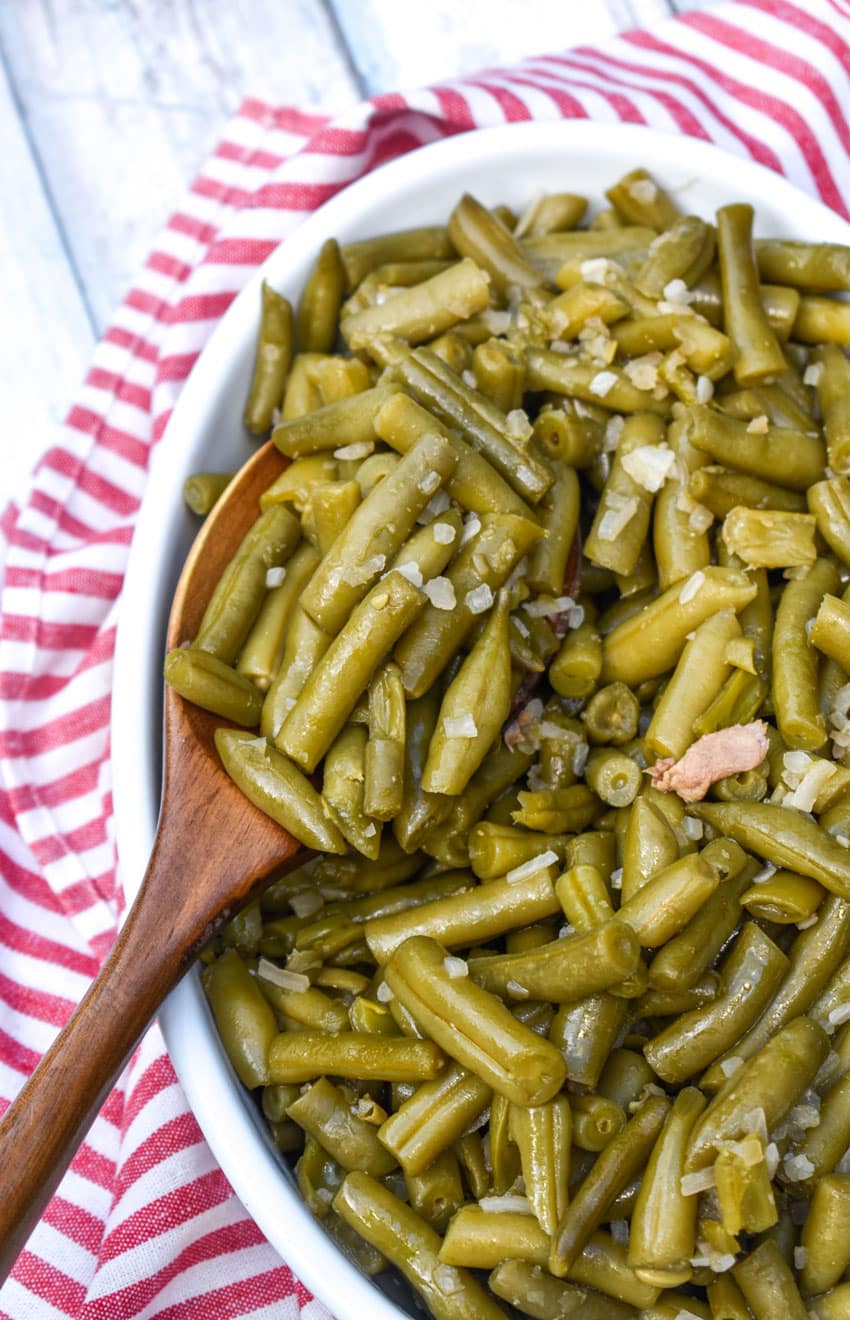 a wooden spoon stuck in slow cooker green beans in a white serving dish