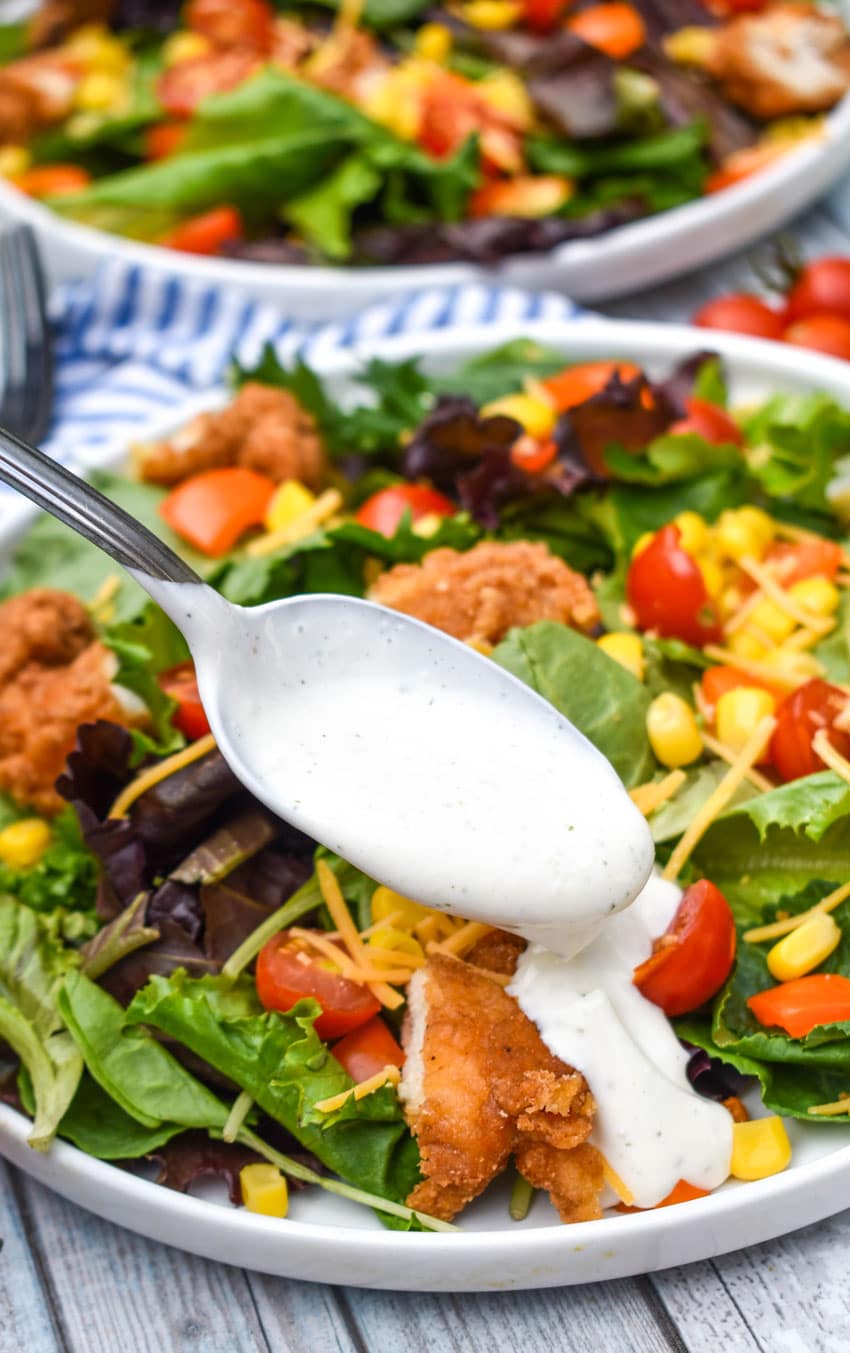 a silver spoon drizzling tiktok cottage cheese ranch dressing over a garden salad on a white plate