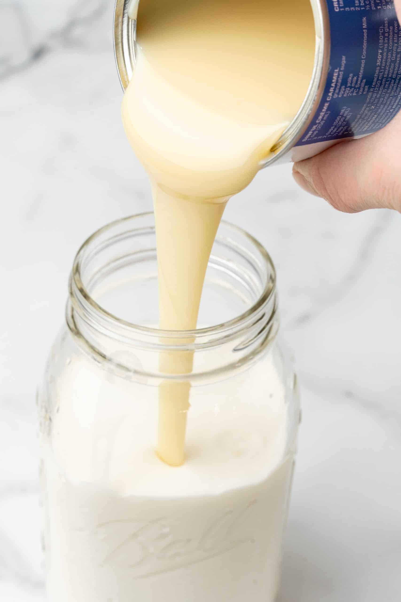 sweetened condensed milk being poured into a glass jar filled with milk