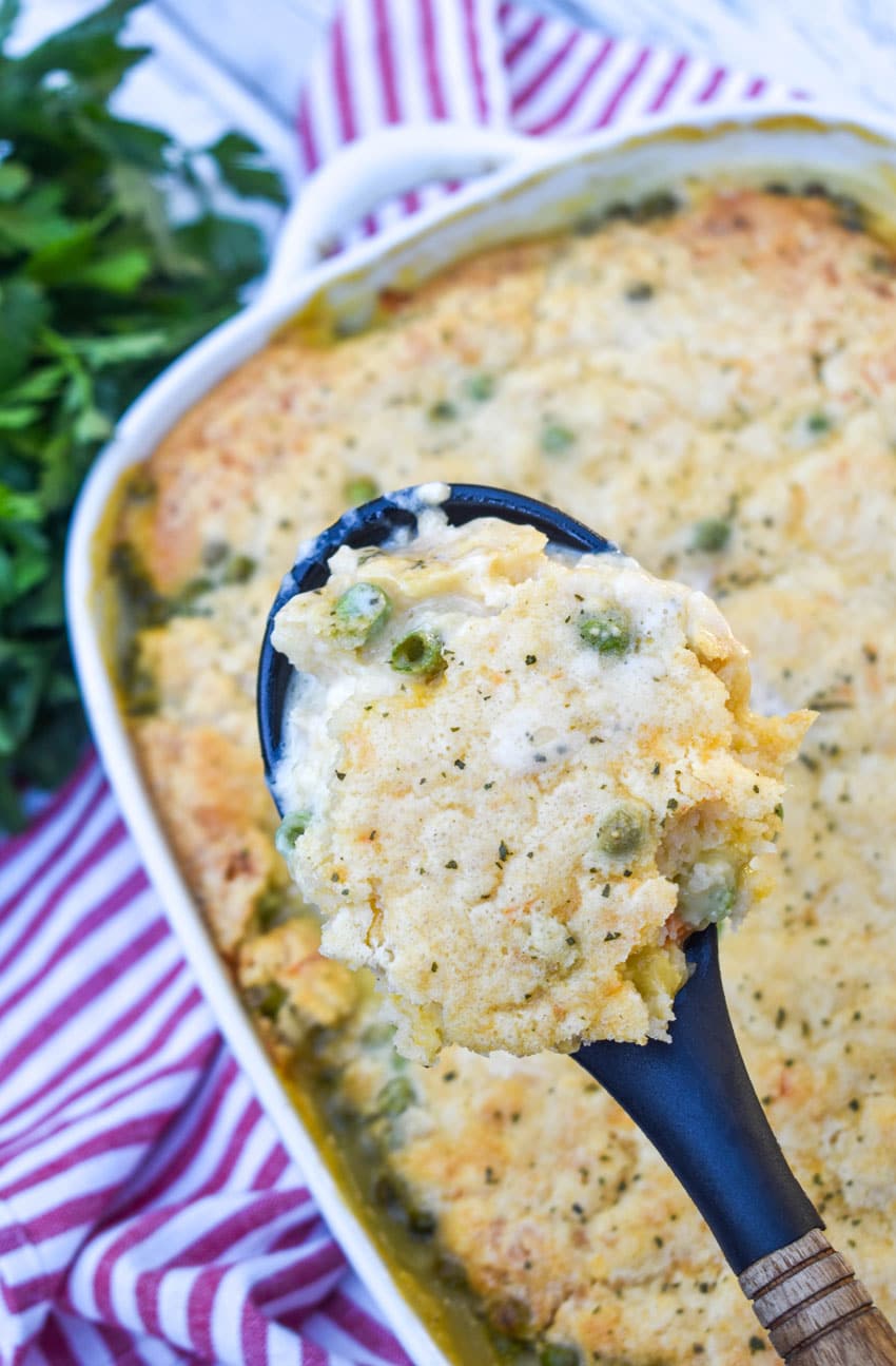a black spoon with a wooden handle holding up a scoop of cheddar bay biscuit chicken cobbler above a white casserole dish