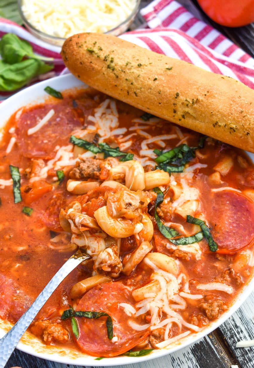 a silver spoon scooping pizza soup out of a white bowl with a bread stick resting on the edge