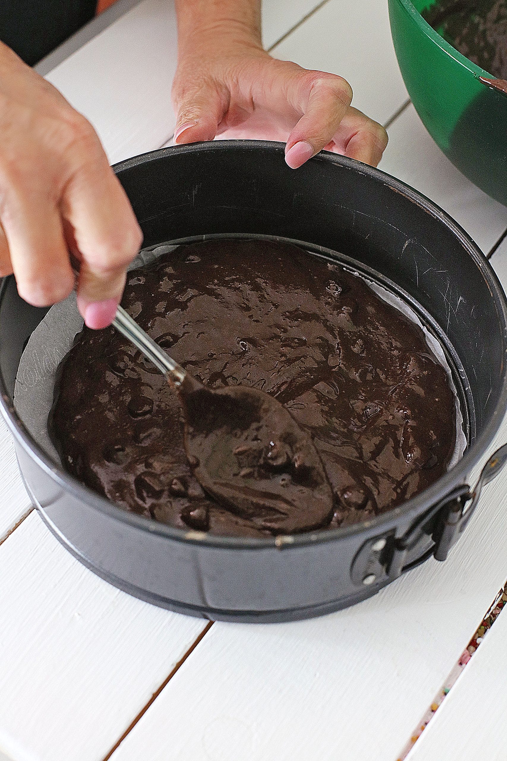 a hand using a silver spoon to spread brownie batter in the the bottom of a metal springform pan