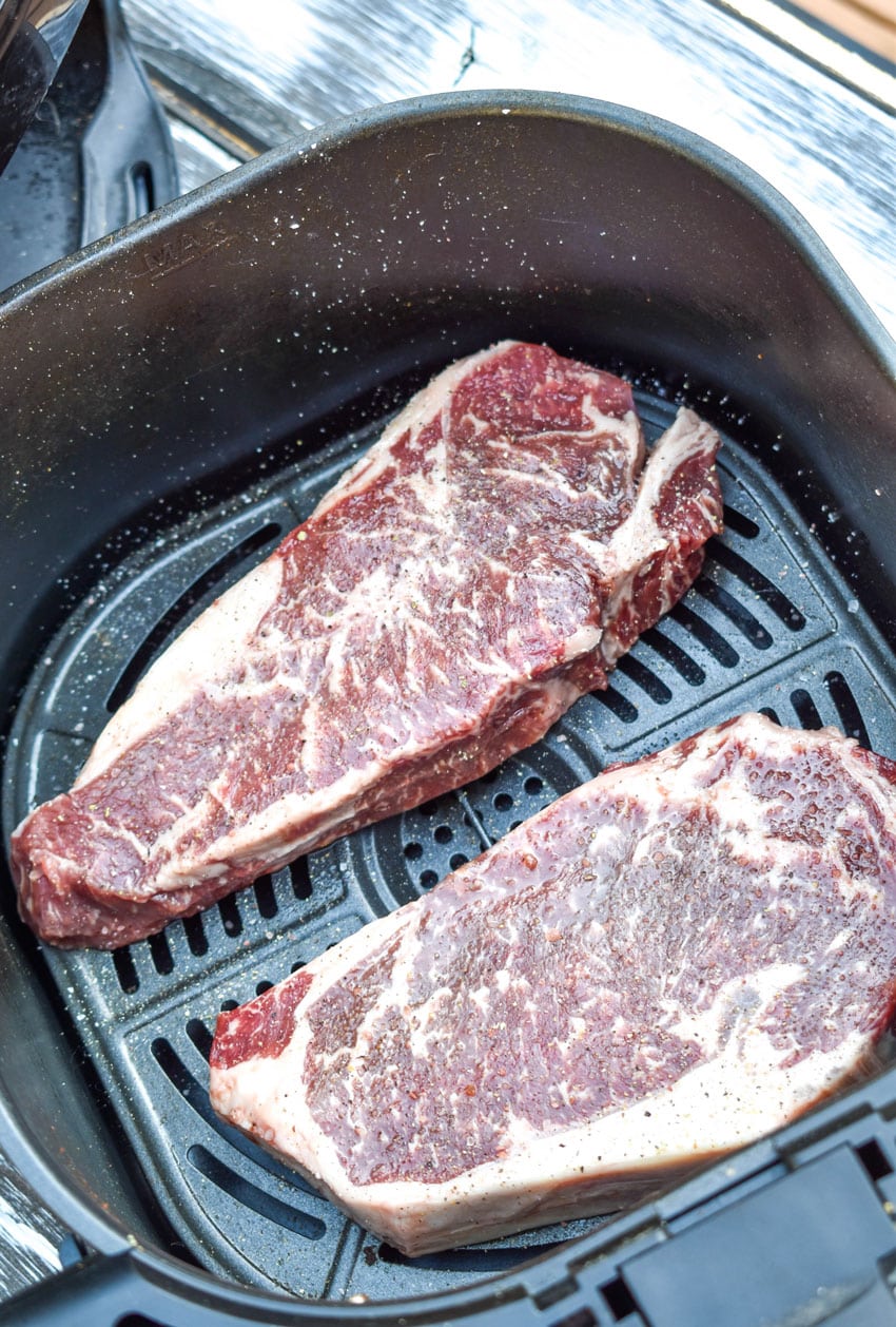 two salted and peppered uncooked steaks in the basket of an air fryer