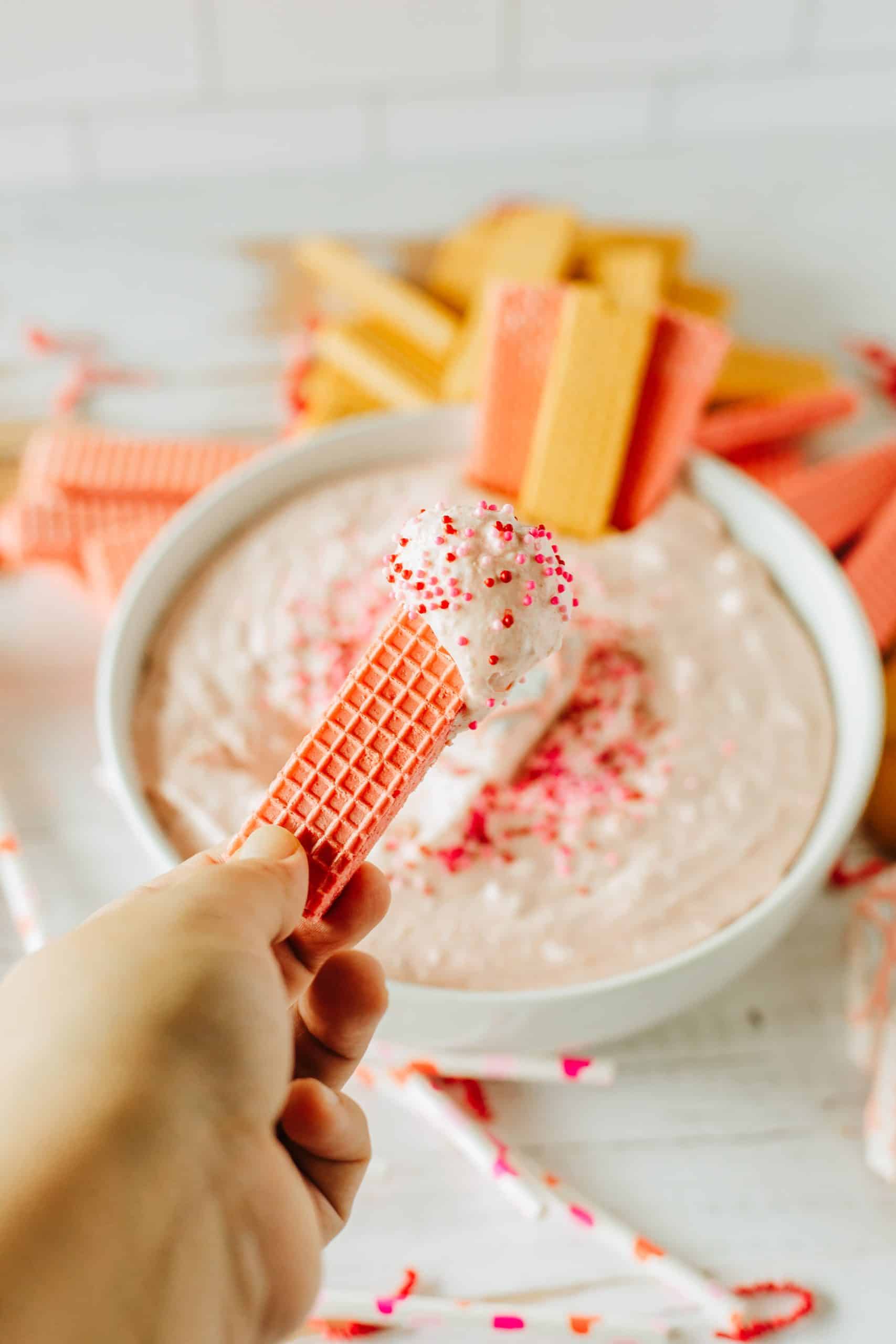 a hand holding up a pink wafer cookie topped with little debbie valentine cake dip
