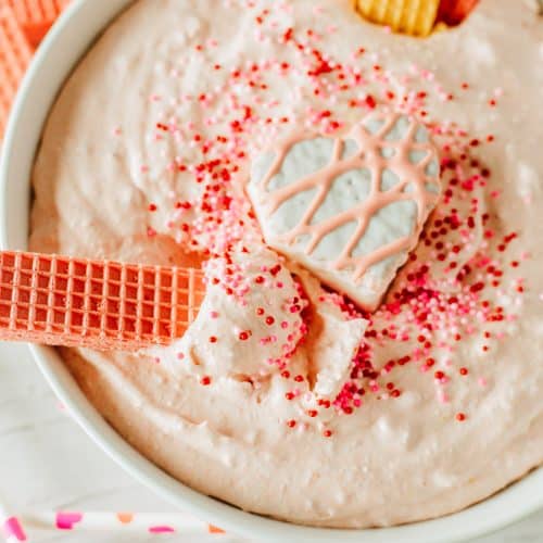 a vanilla wafer cookie stick in a bowl of Little Debbie Valentine cake dip in a white bowl topped with sprinkles and a snack cake