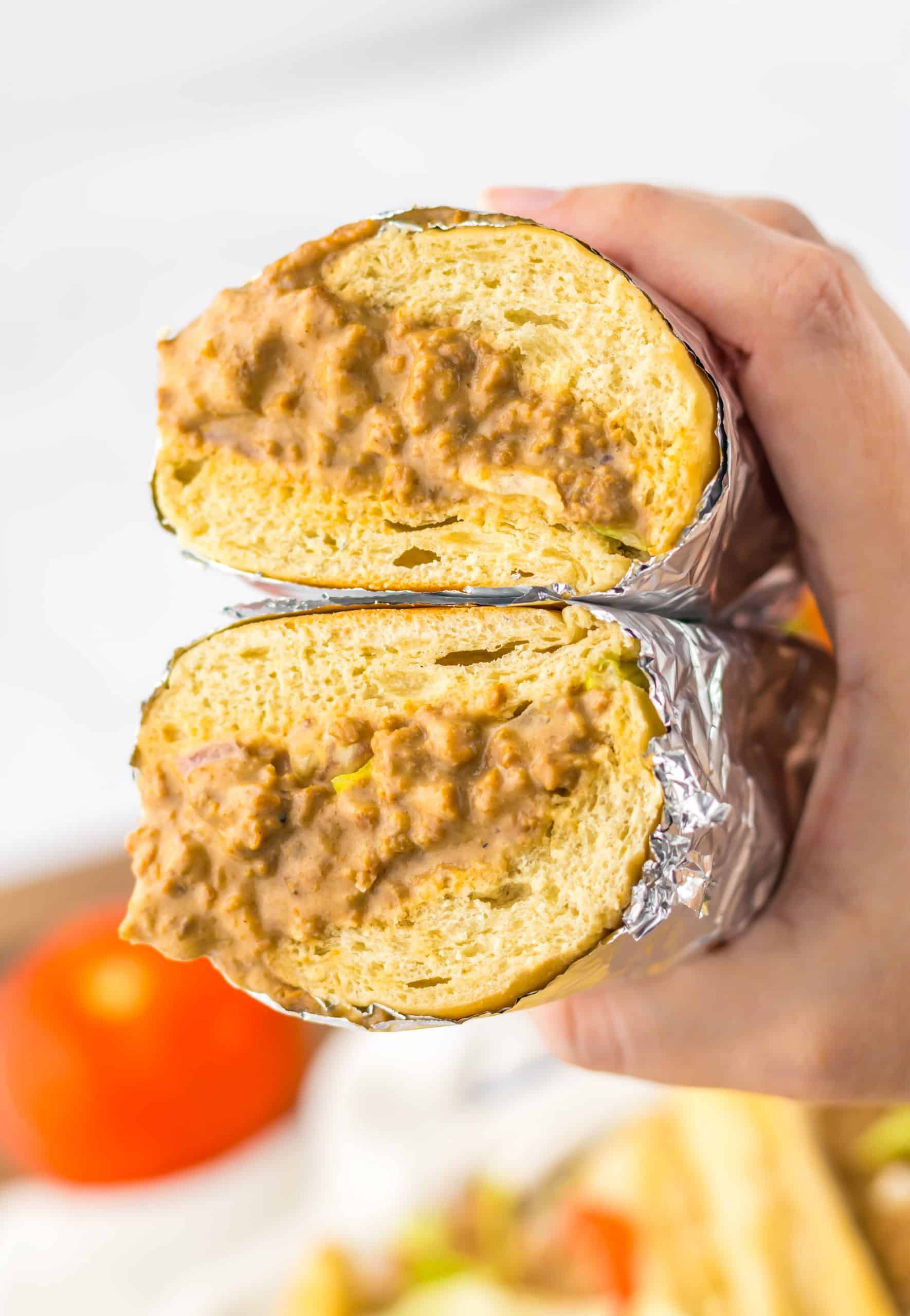 a hand holding up two halves of a chopped cheeseburger sub wrapped in tin foil