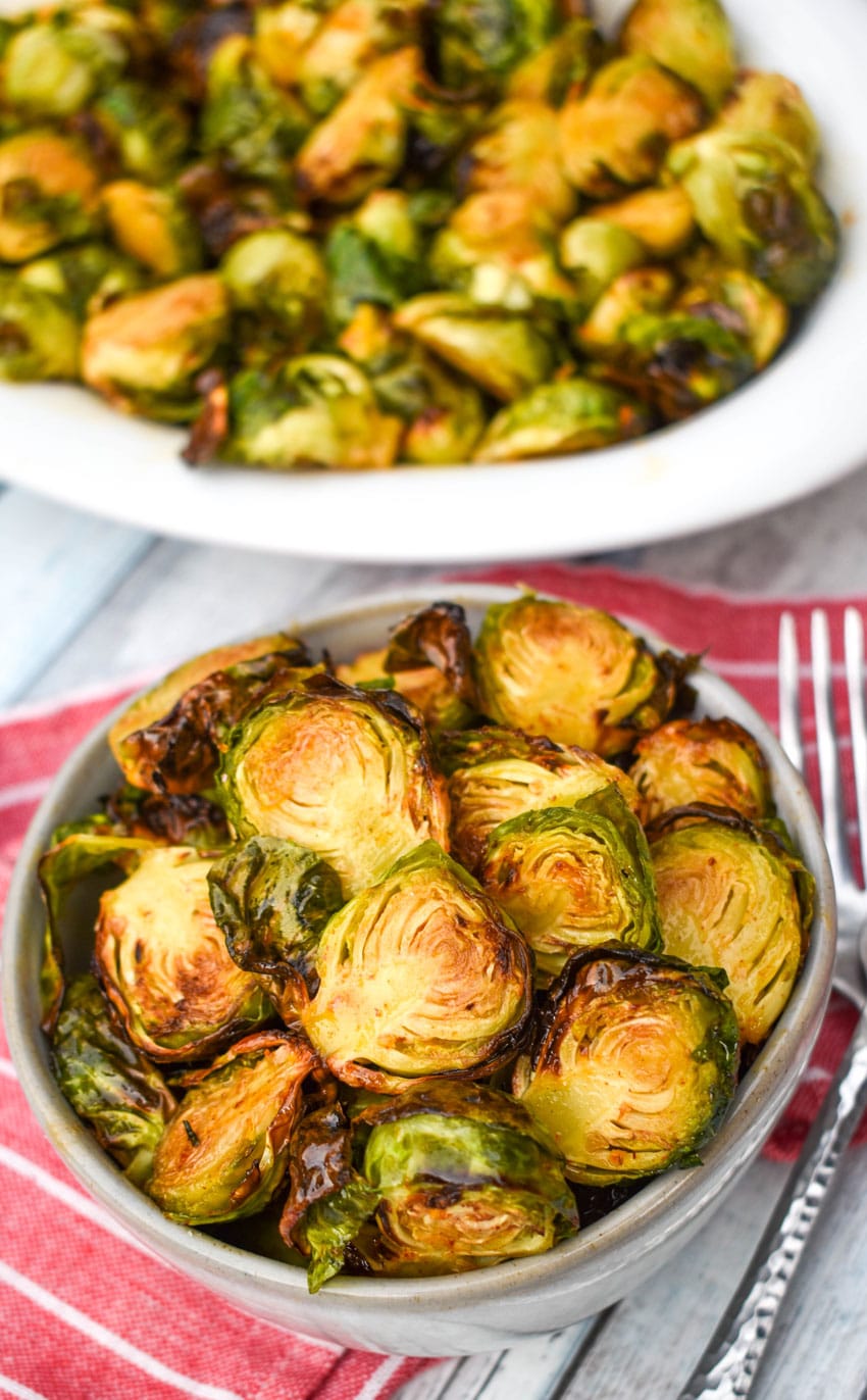 glazed air fried brussels sprouts in a small gray bowl in front of a white serving dish with a silver fork on the side