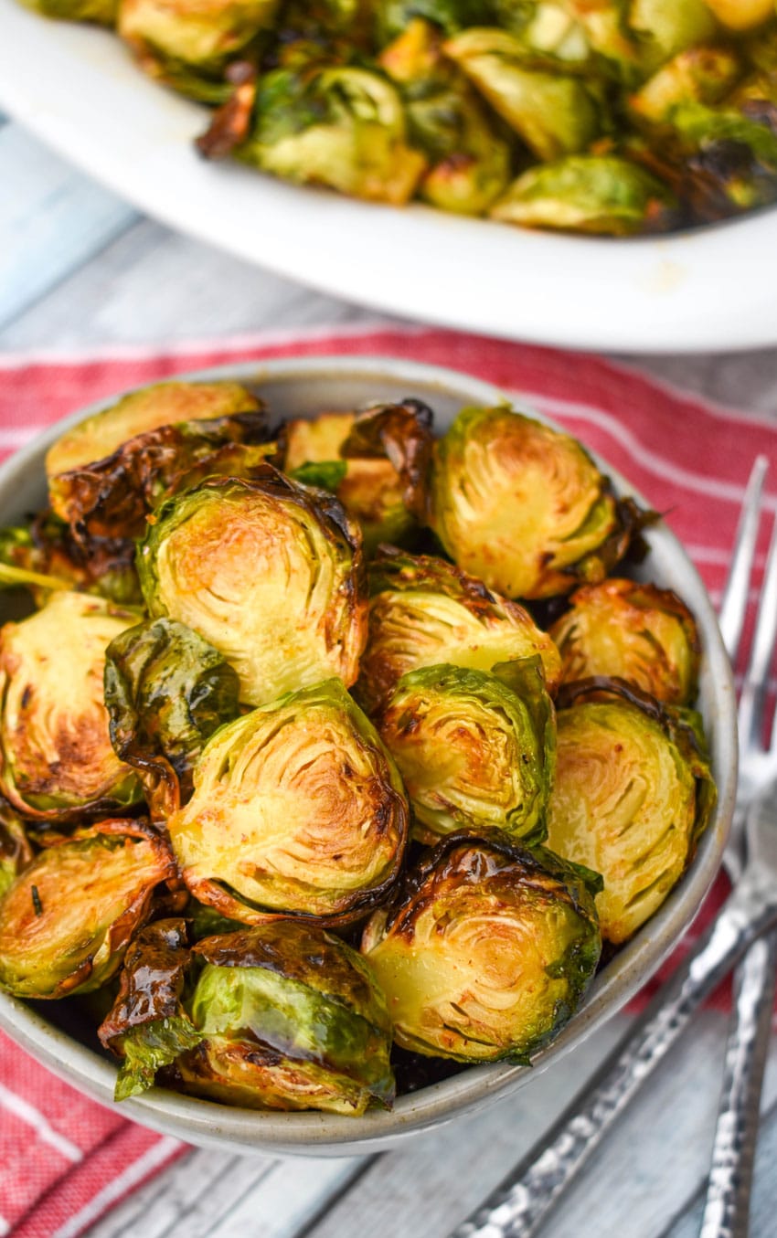 glazed air fried brussels sprouts in a small gray bowl in front of a white serving dish with a silver fork on the side