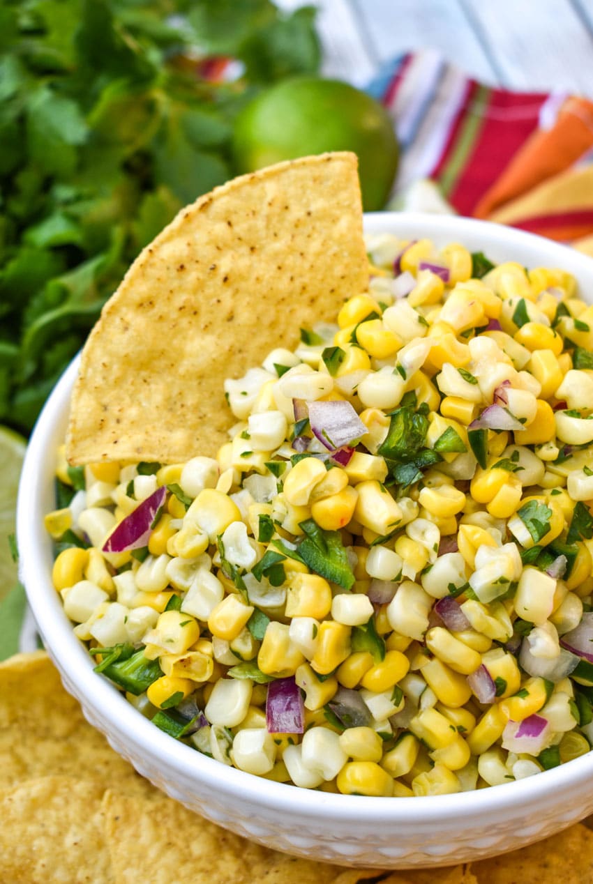 a fresh corn salsa recipe in a white bowl with a tortilla chip stuck on top