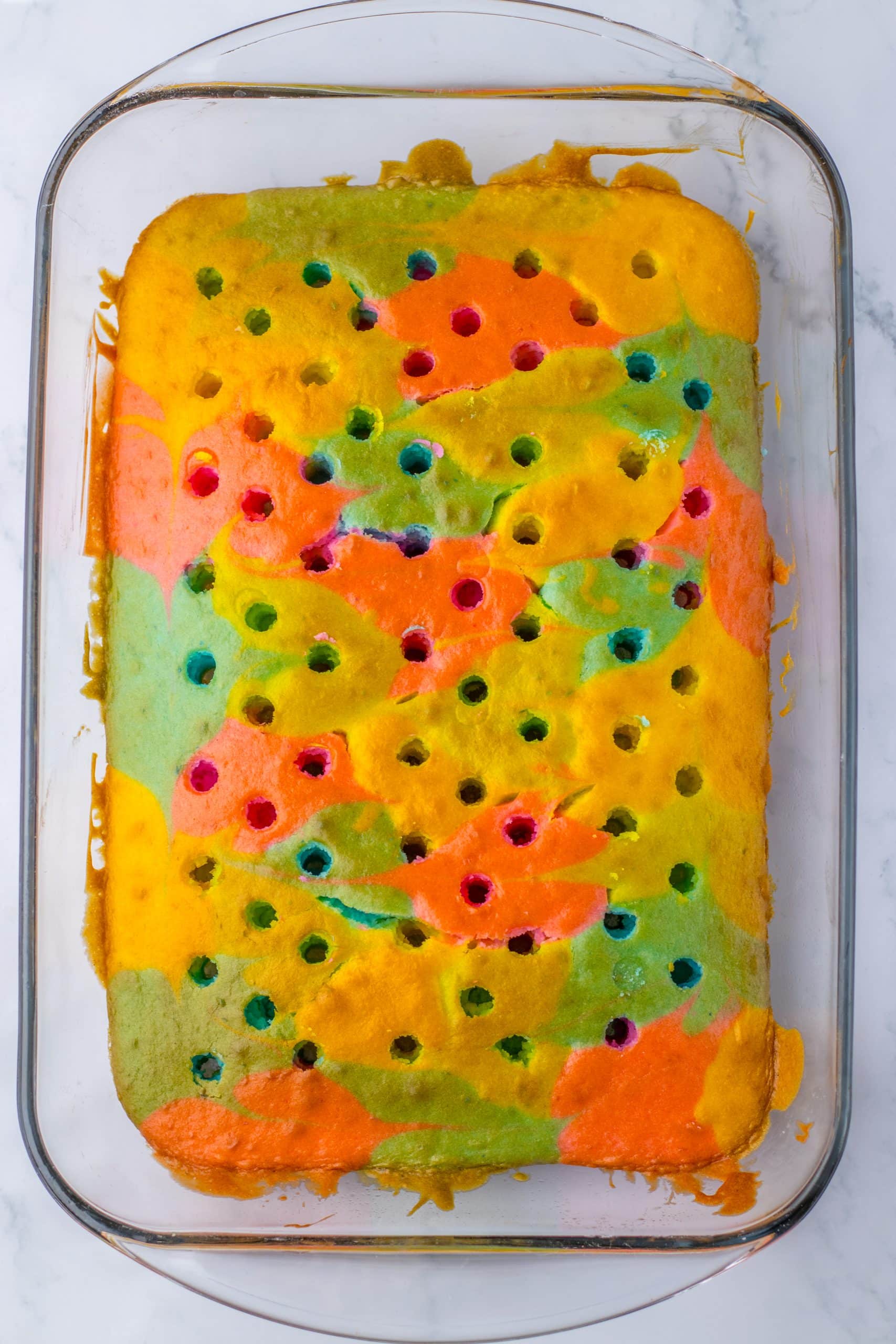 a rainbow colored cake with holes poked in the top in a rectangular glass baking dish
