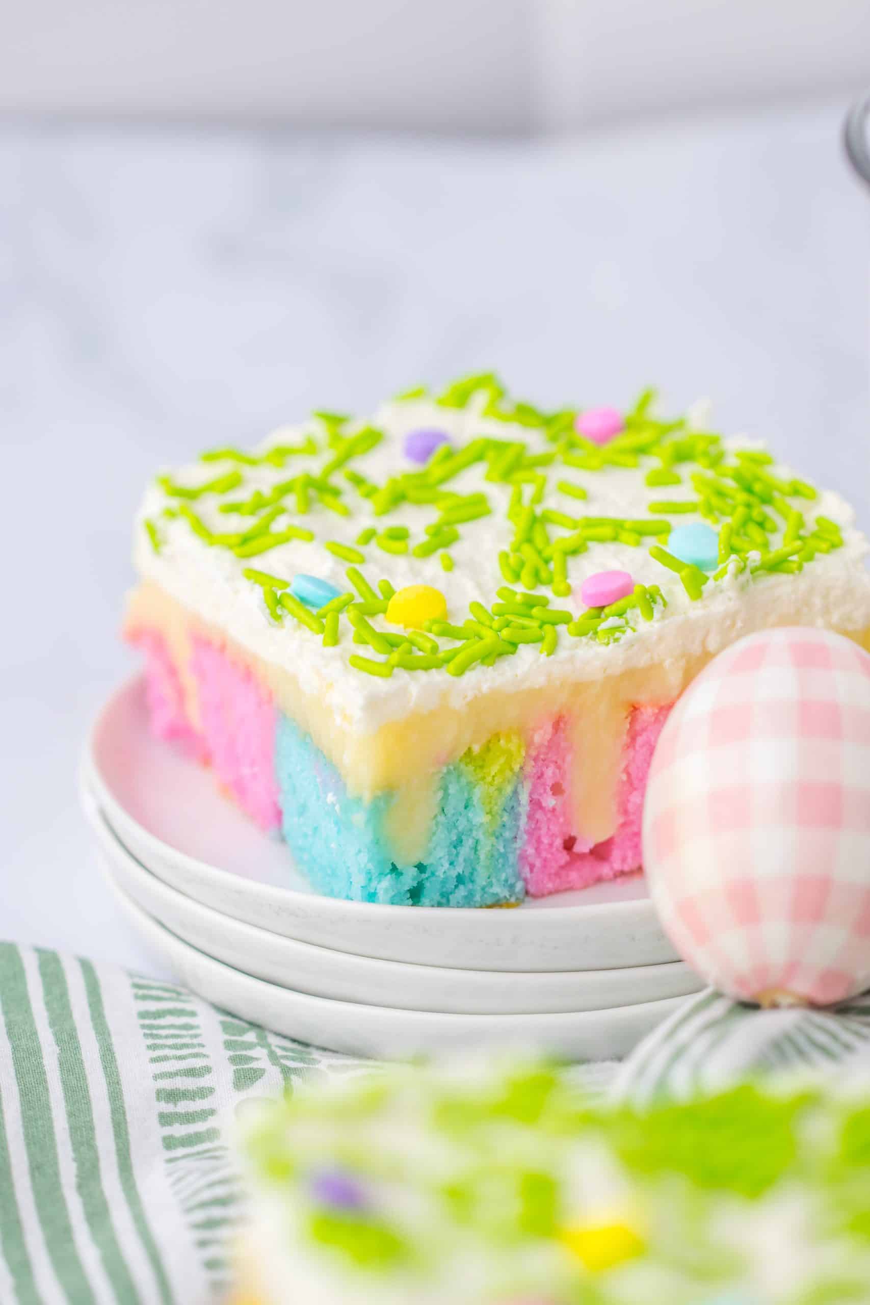 Easter Poke Cake a slice of pastel rainbow colored easter poke cake on a small white dessert plate with a glass of milk in the background