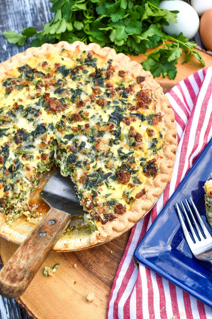 a wooden handled spatula resting in a sliced sausage and spinach quiche on a wooden cutting board