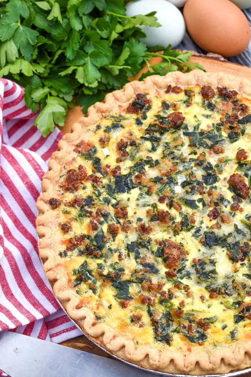 a sausage and spinach quiche on a wooden cutting boar with fresh green herbs in the background