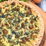 a sausage and spinach quiche on a wooden cutting boar with fresh green herbs in the background