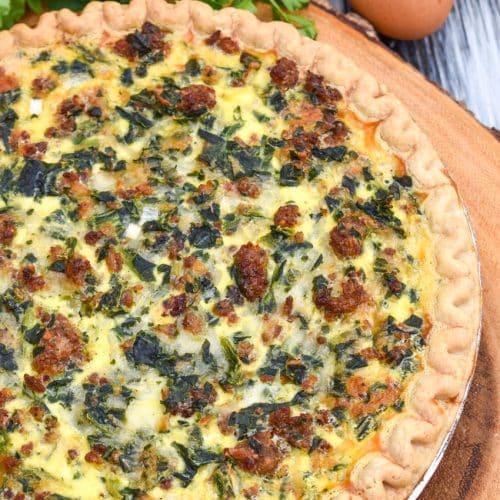 a sausage and spinach quiche on a wooden cutting boar with fresh green herbs in the background