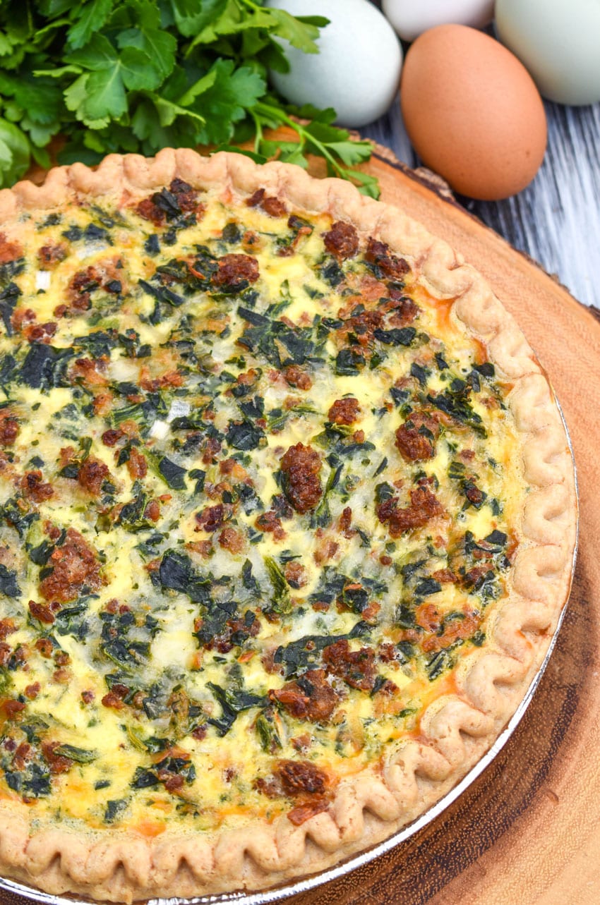 a sausage and spinach quiche on a wooden cutting boar with fresh green herbs in the background