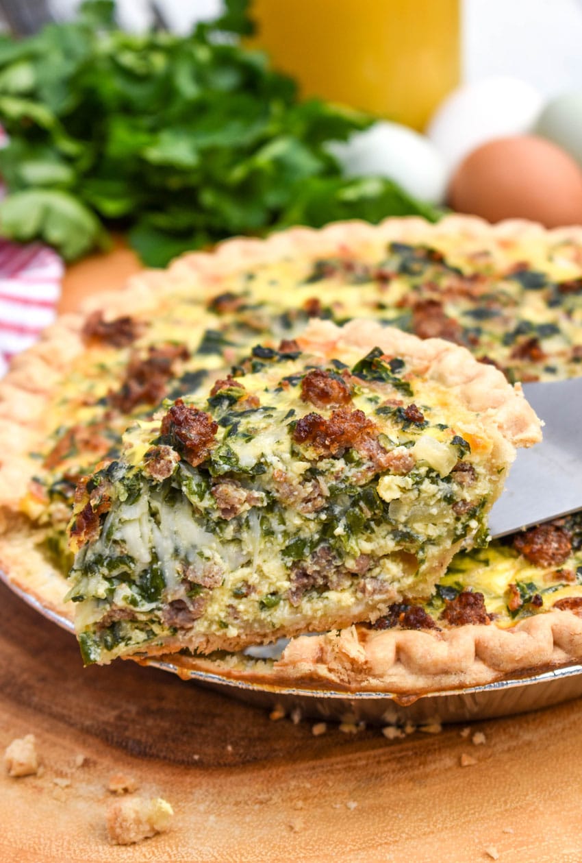 a wooden handled pie server holding a slice of spinach and sausage quiche resting on the edge of the whole pie