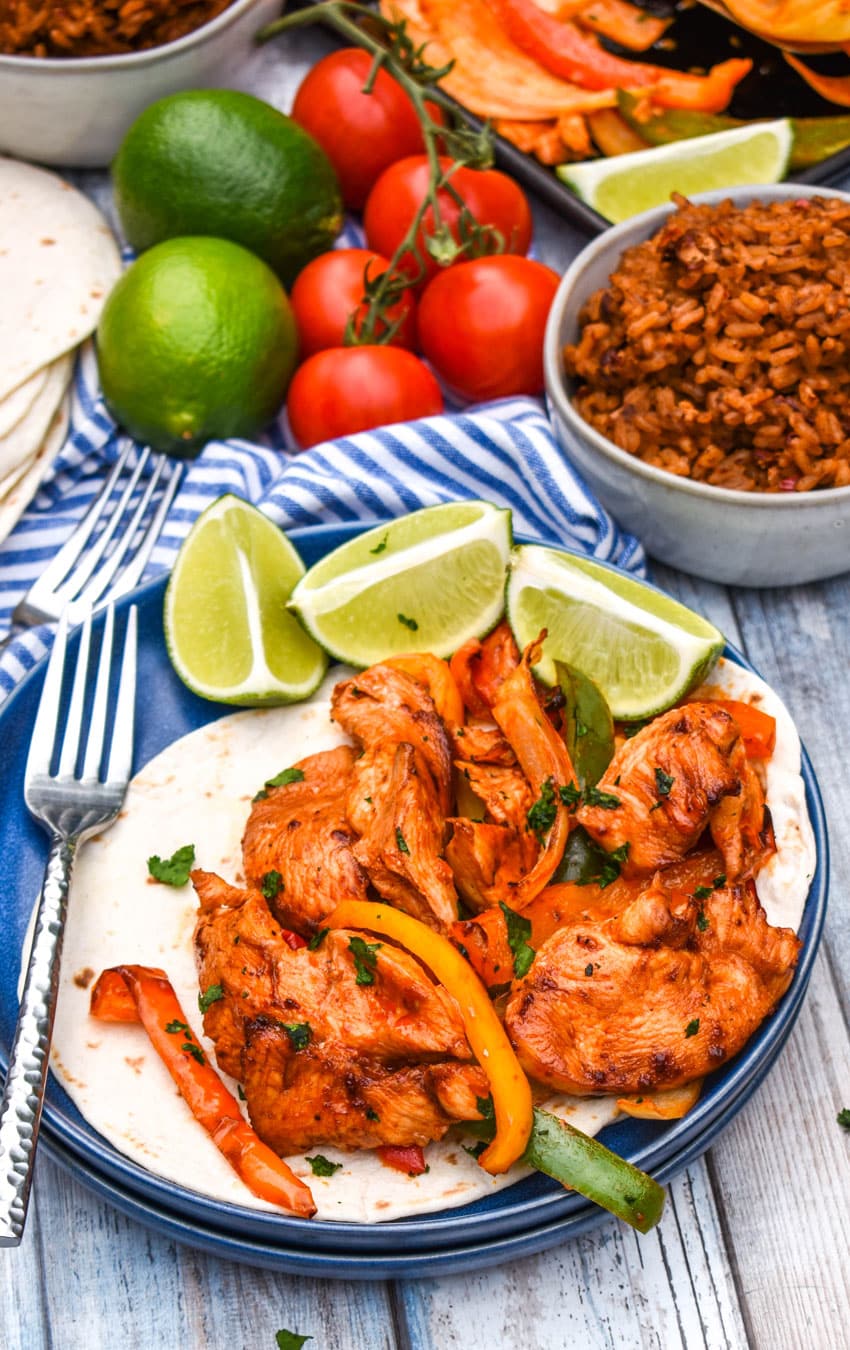 sheet pan chicken fajitas mixture on flour tortillas on a blue plate with fresh lime wedges on the side