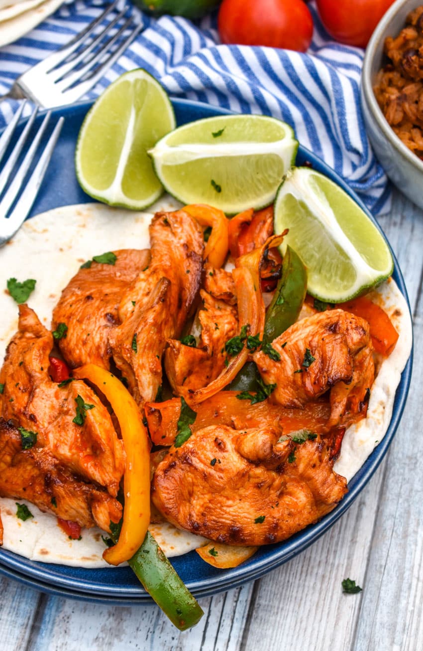 sheet pan chicken fajitas mixture on flour tortillas on a blue plate with fresh lime wedges on the side