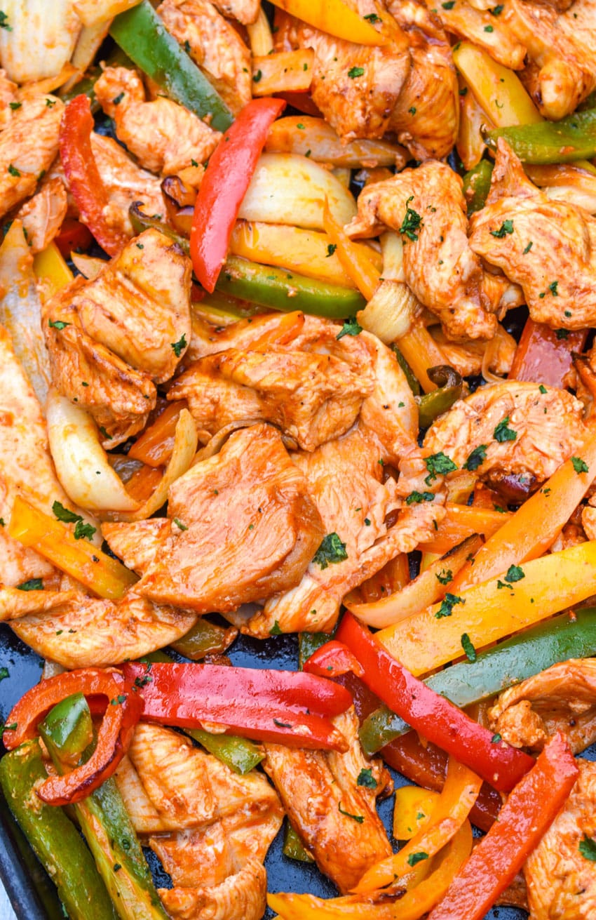 a close up of chicken and sliced veggies in fajita seasoning on a metal sheet pan