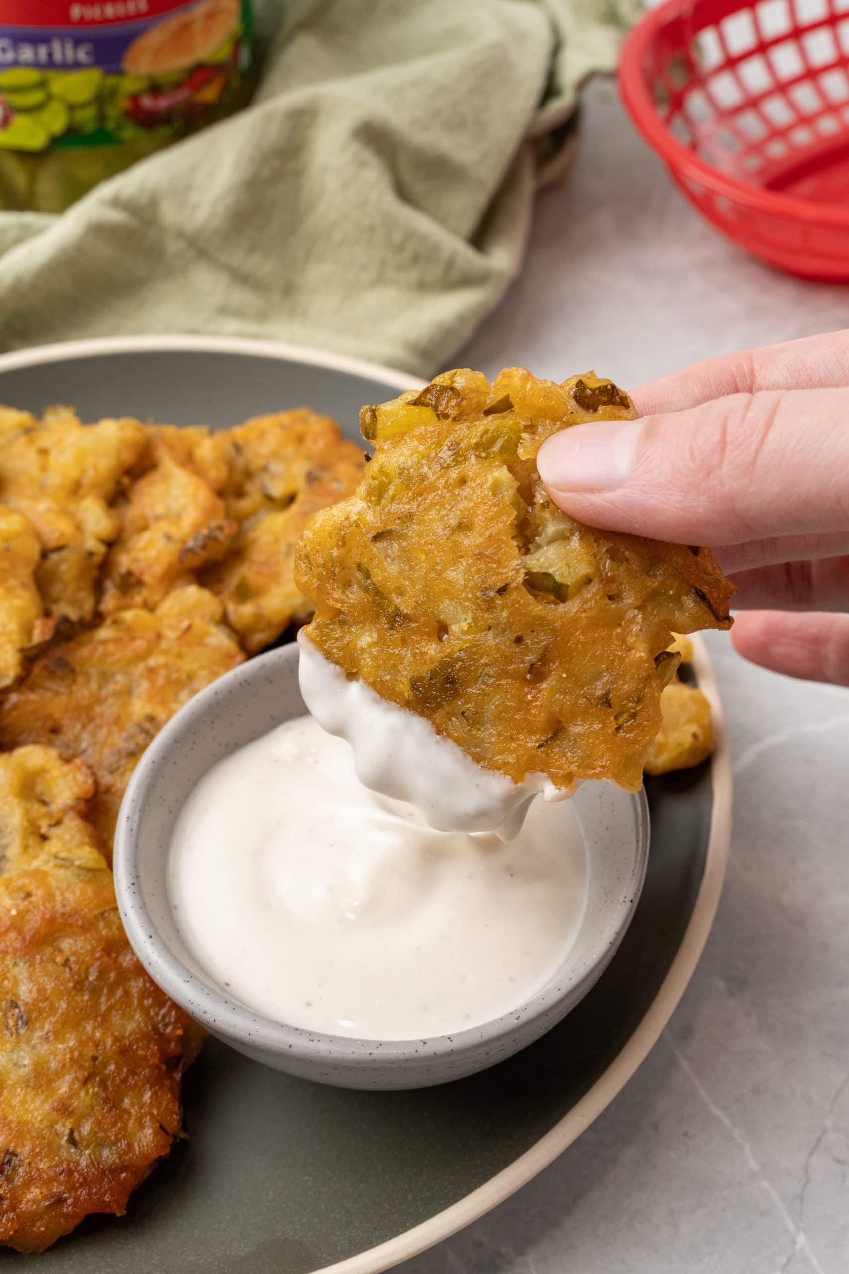 a hand holding a dill pickle fritter being dipped into a small gray bowl filled with ranch dressing