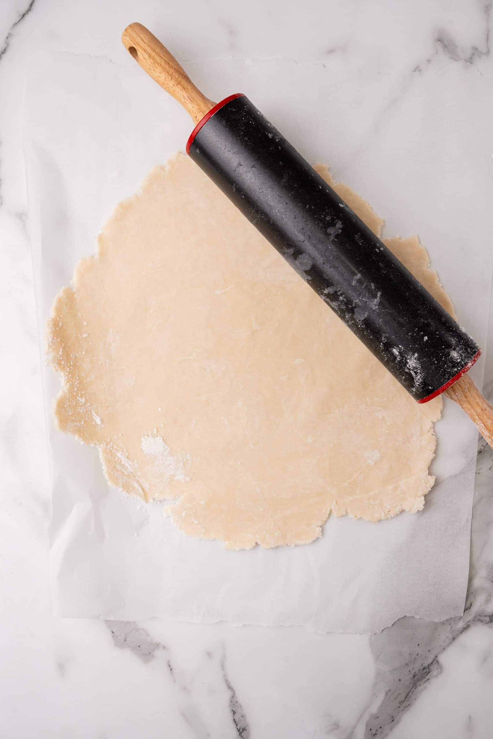 an easy homemade pie crust rolled out on a white piece of parchment paper with a black rolling pin resting on the side