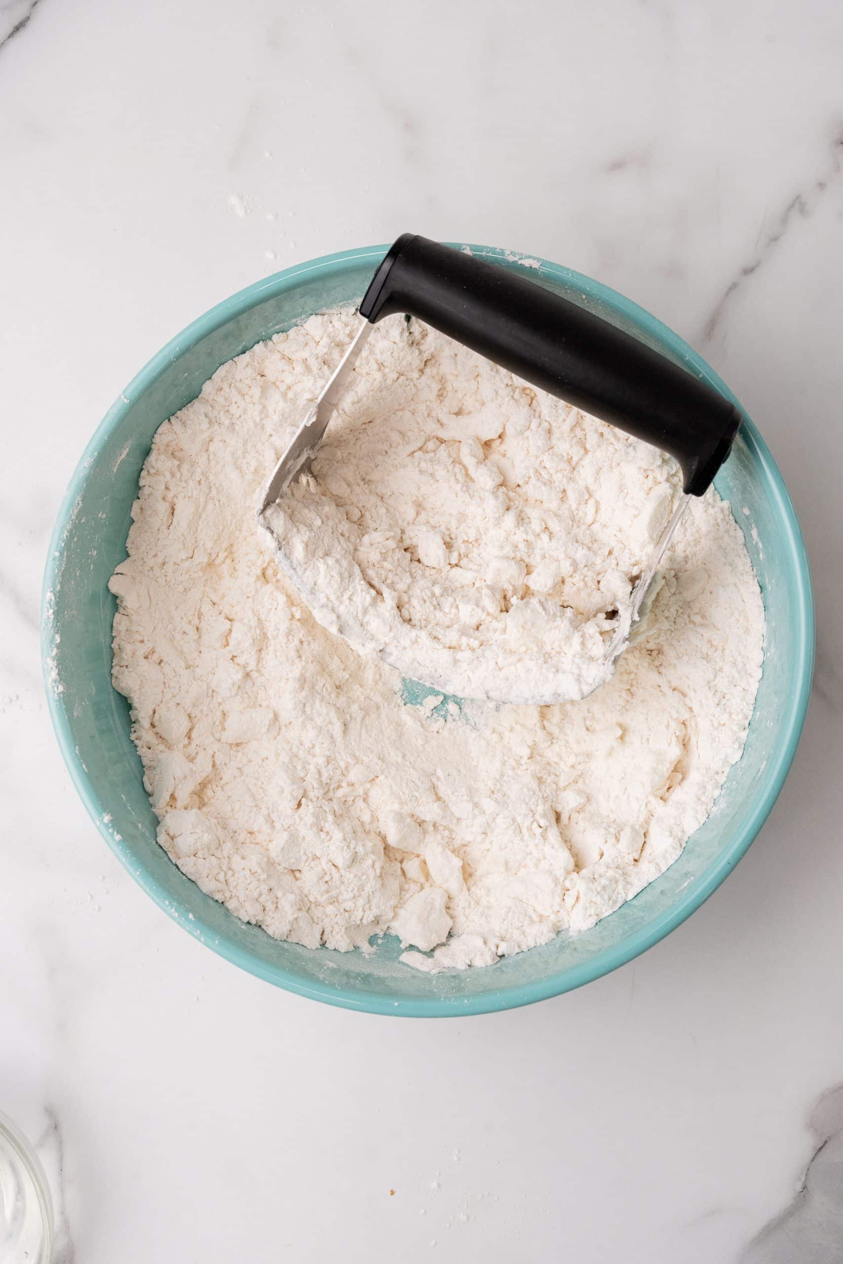 a pastry blender resting on the edge of a blue mixing bowl filled with flour and lard