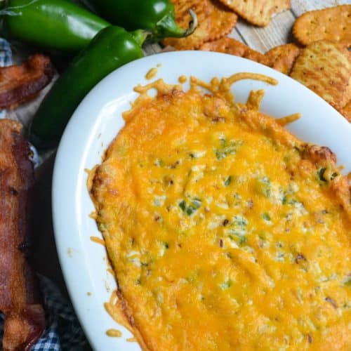 jalapeno popper di in a white serving dish surrounded by strips of crisp bacon, jalapeno peppers, and pretzel crackers