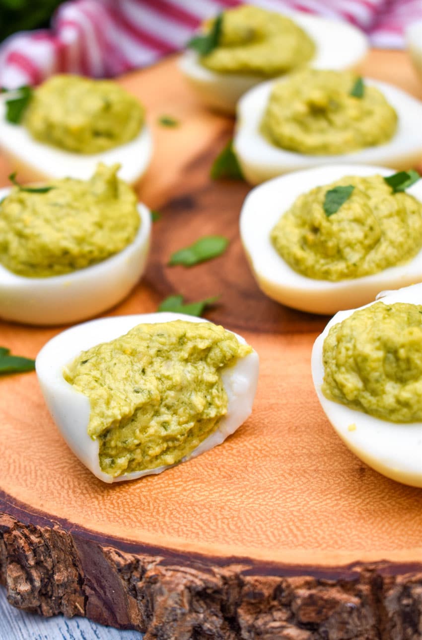 pesto deviled eggs topped with fresh parsley arranged on a wooden cutting board with the front egg bitten to reveal the filling inside