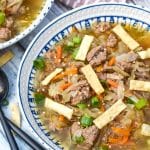 pork egg roll soup topped with crispy wonton strips in a white bowl with two black spoons on the side