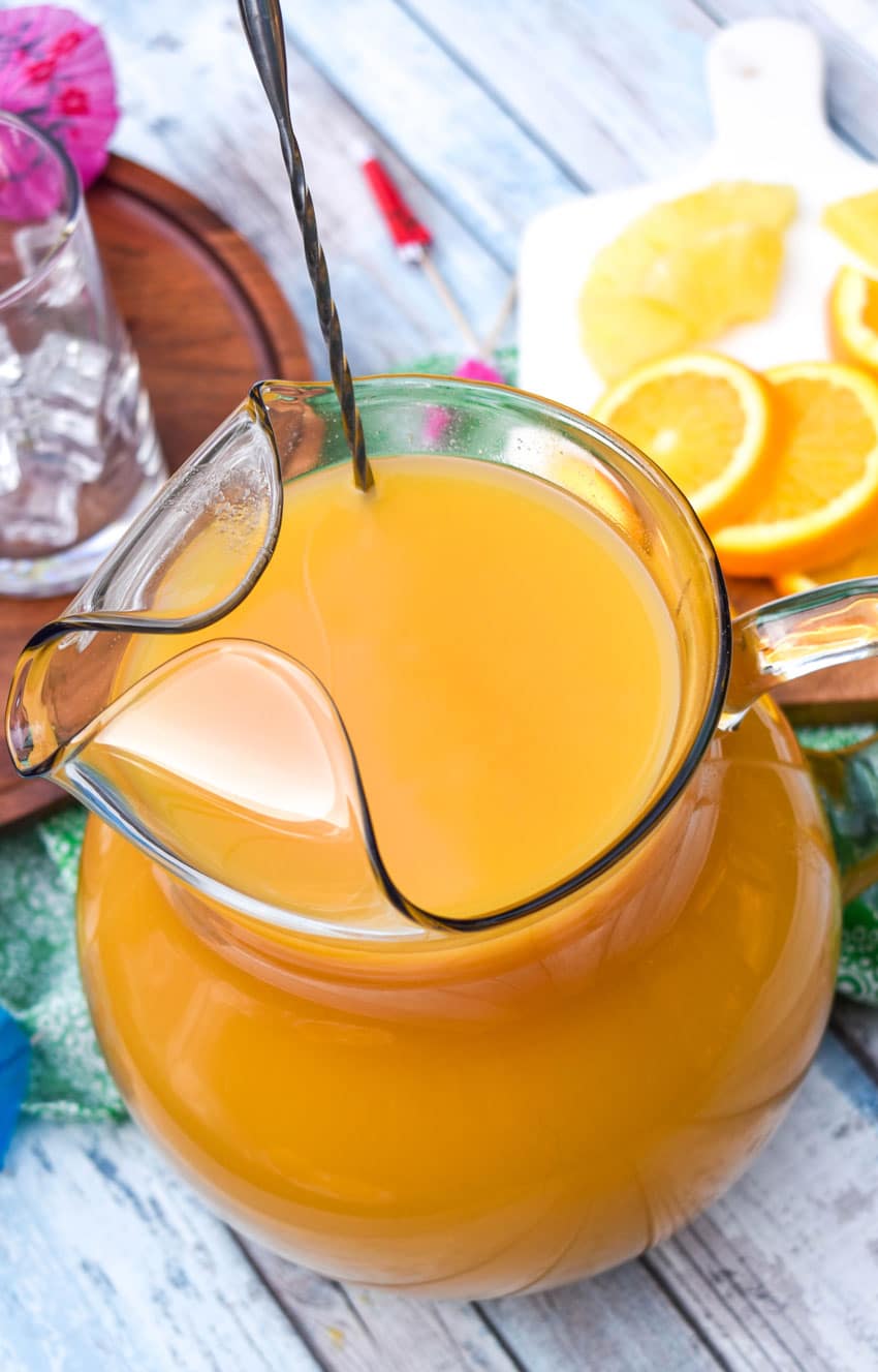 disney pog juice in a glass pitcher with a metal stir stick resting against the edge