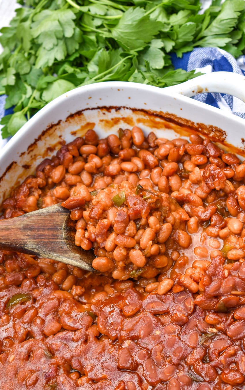 a wooden spoon scooping the best canned baked beans out of a white casserole dish