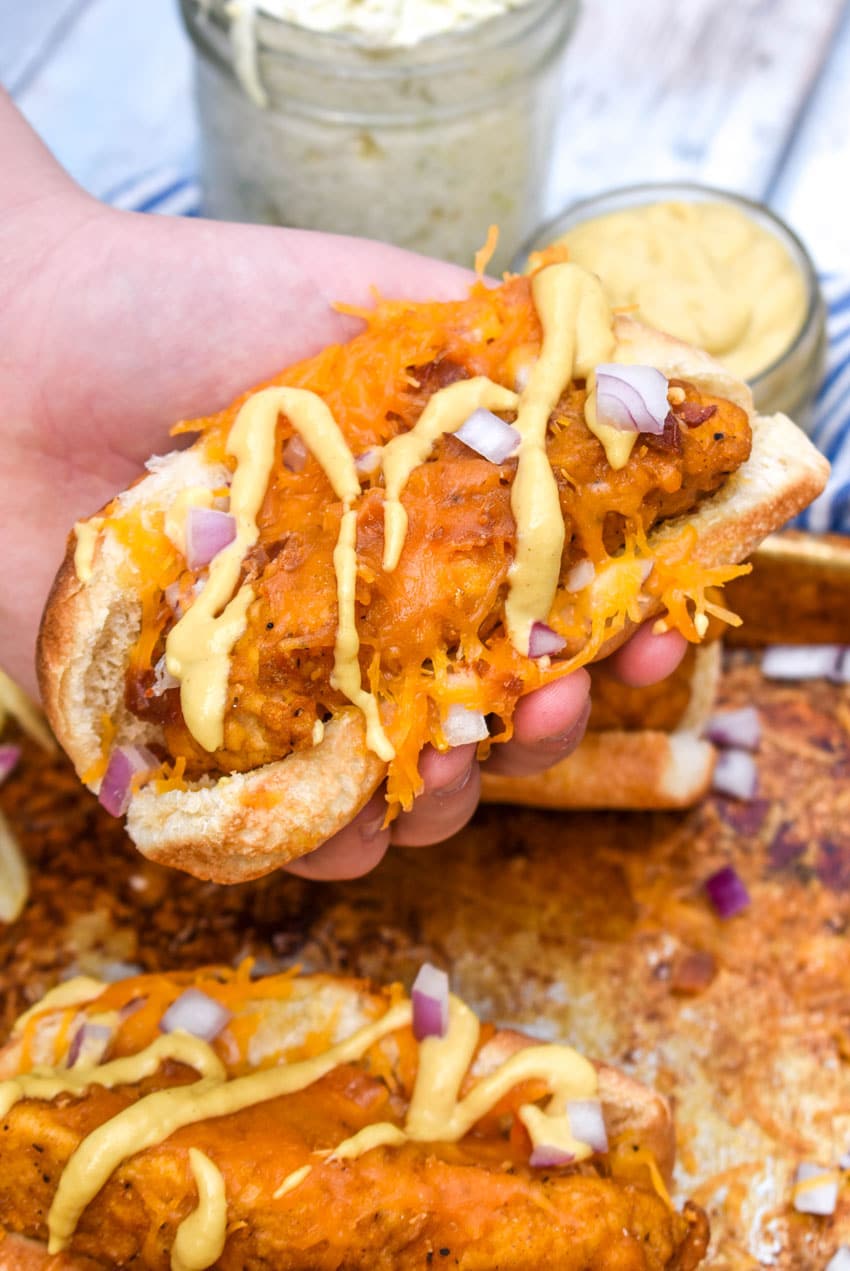 a hand holding up a southern bird dog sandwich topped with drizzled honey mustard and diced red onion