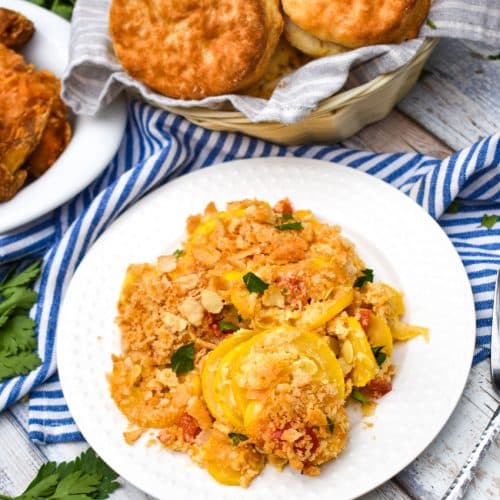 southern squash casserole on a small white plate with a basket of biscuits and platter of fried chicken on the side