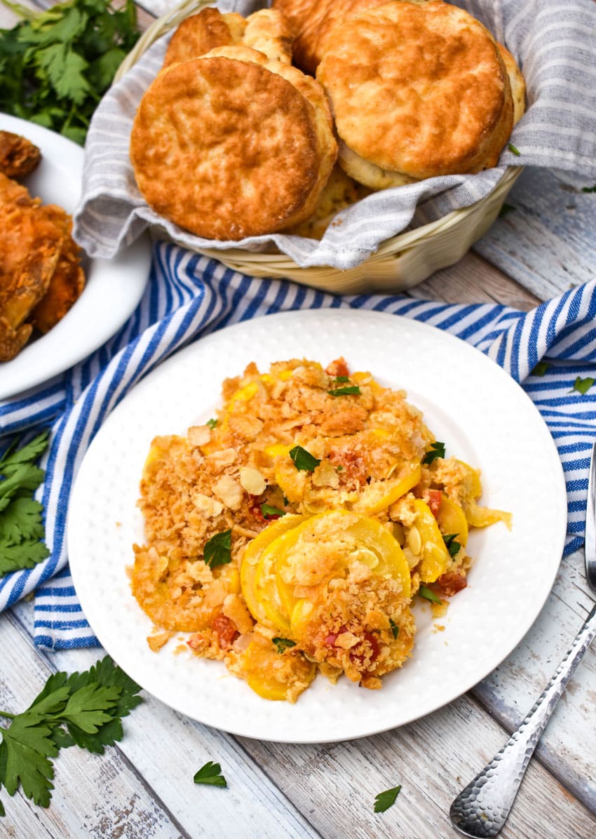 southern squash casserole on a small white plate with a basket of biscuits and platter of fried chicken on the side