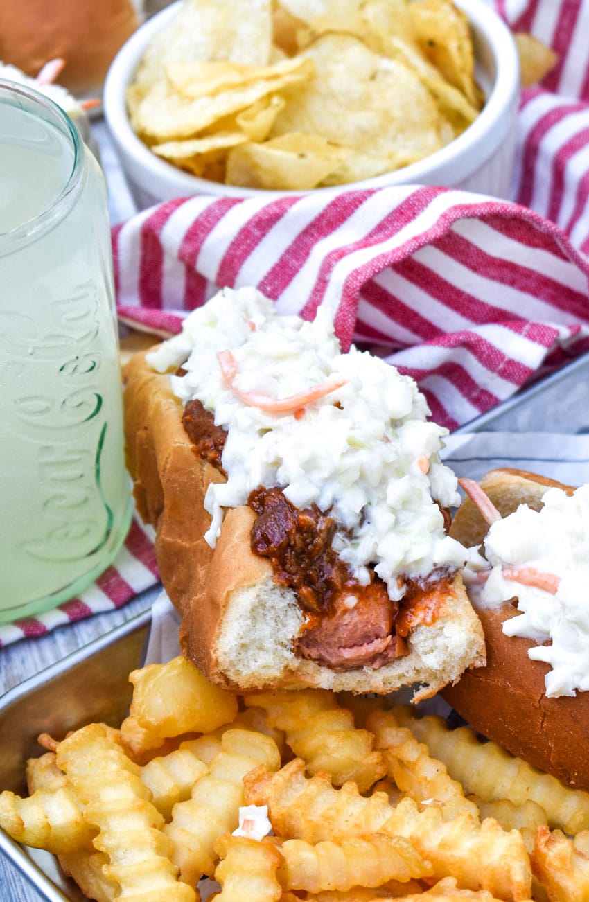 a bitten southern slaw dog resting on the edge of a metal serving tray holding crispy crinkle cut french fries 