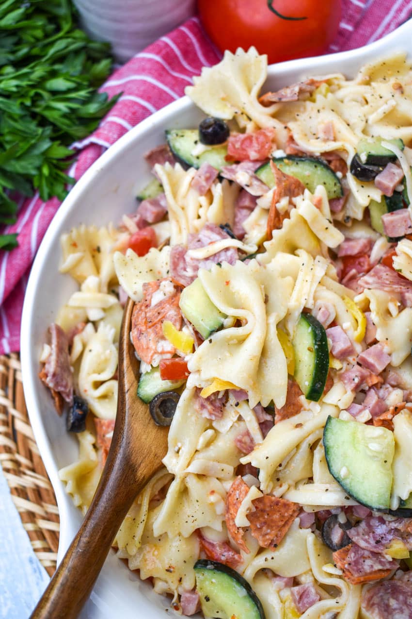 a closeup of a wooden spoon resting in a white bowl filled with italian pasta salad