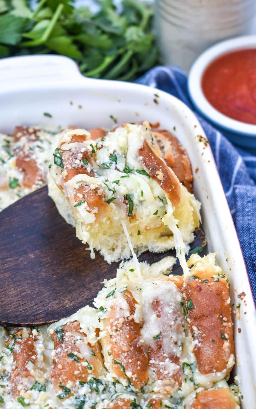 a wooden spatula lifting a single piece of cheesy hawaiian roll garlic bread out of a small white baking dish