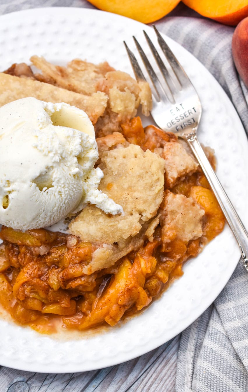a scoop of quick and easy homemade peach cobbler topped with melting vanilla ice cream on a small white plate with a metal fork on the side