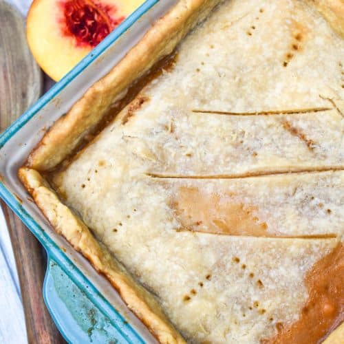 an easy southern peach cobbler with a single crust in a small blue casserole dish with halved fresh peaches on the side