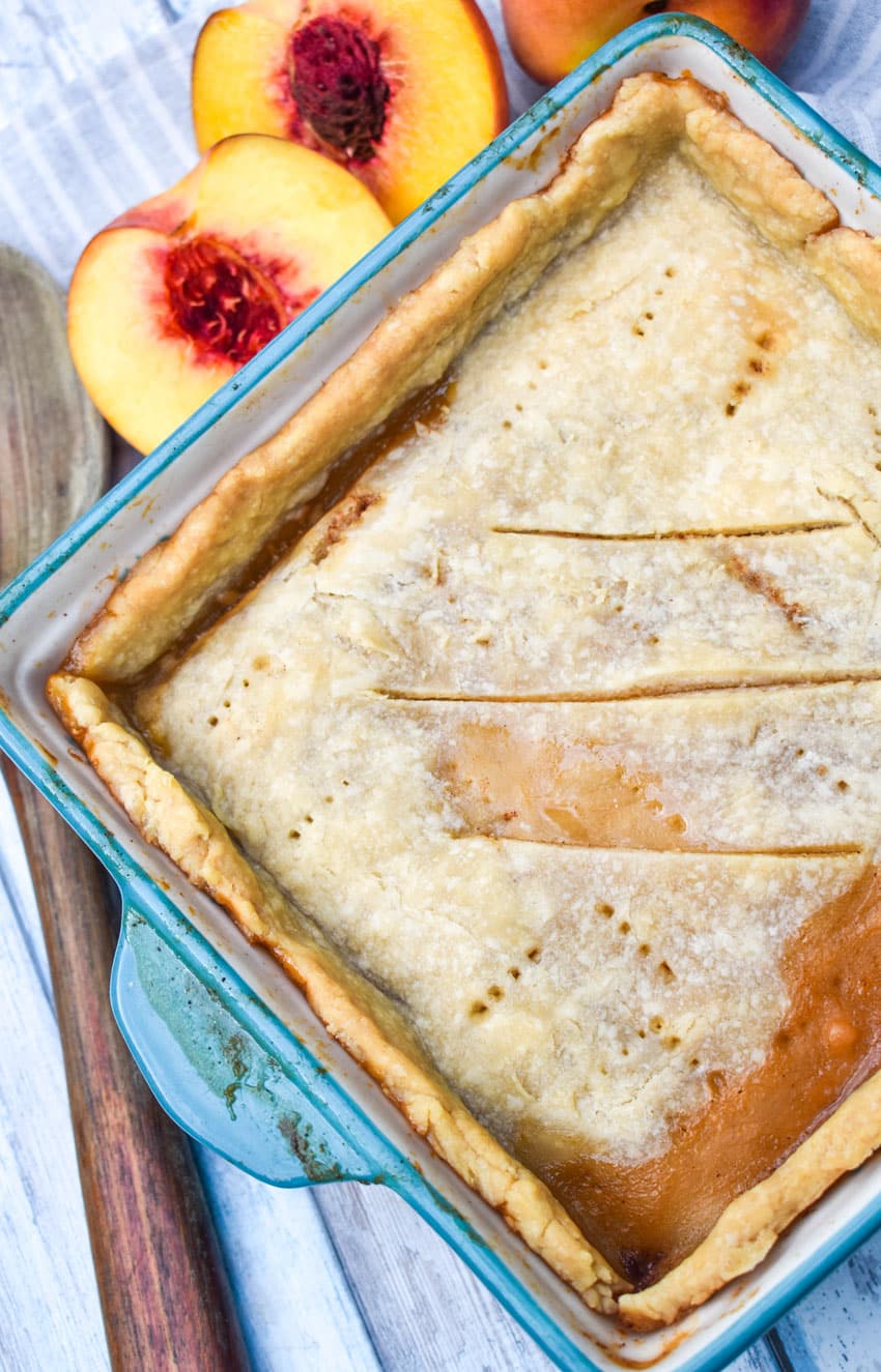 an easy southern peach cobbler with a single crust in a small blue casserole dish with halved fresh peaches on the side