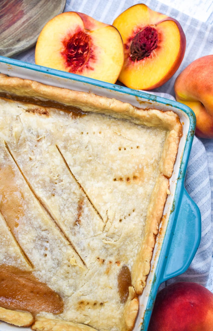 an easy southern peach cobbler with a single crust in a small blue casserole dish with halved fresh peaches on the side
