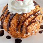 fried ice cream topped with a drizzle of chocolate sauce, whipped cream, and a a maraschino cherry on a small white plate