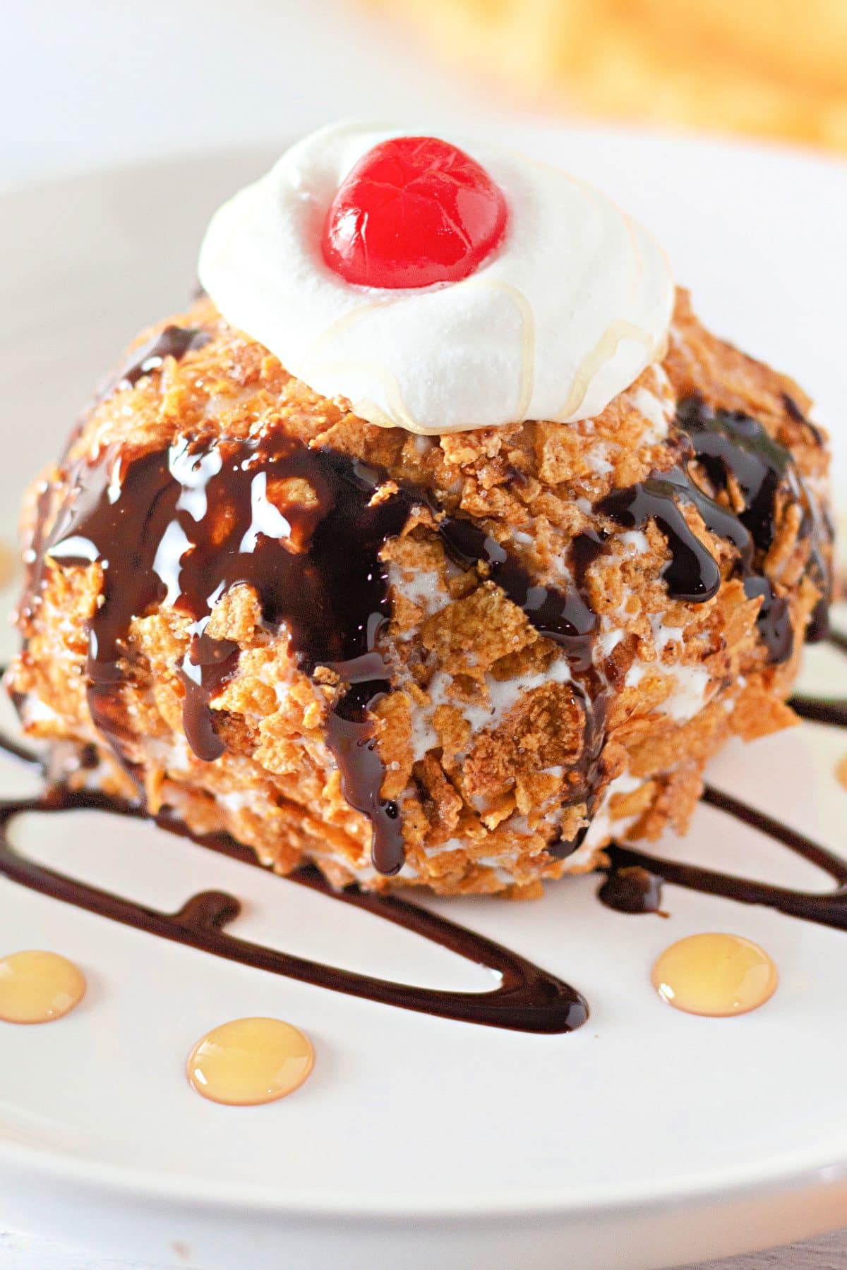 fried ice cream topped with a drizzle of chocolate sauce, whipped cream, and a a maraschino cherry on a small white plate
