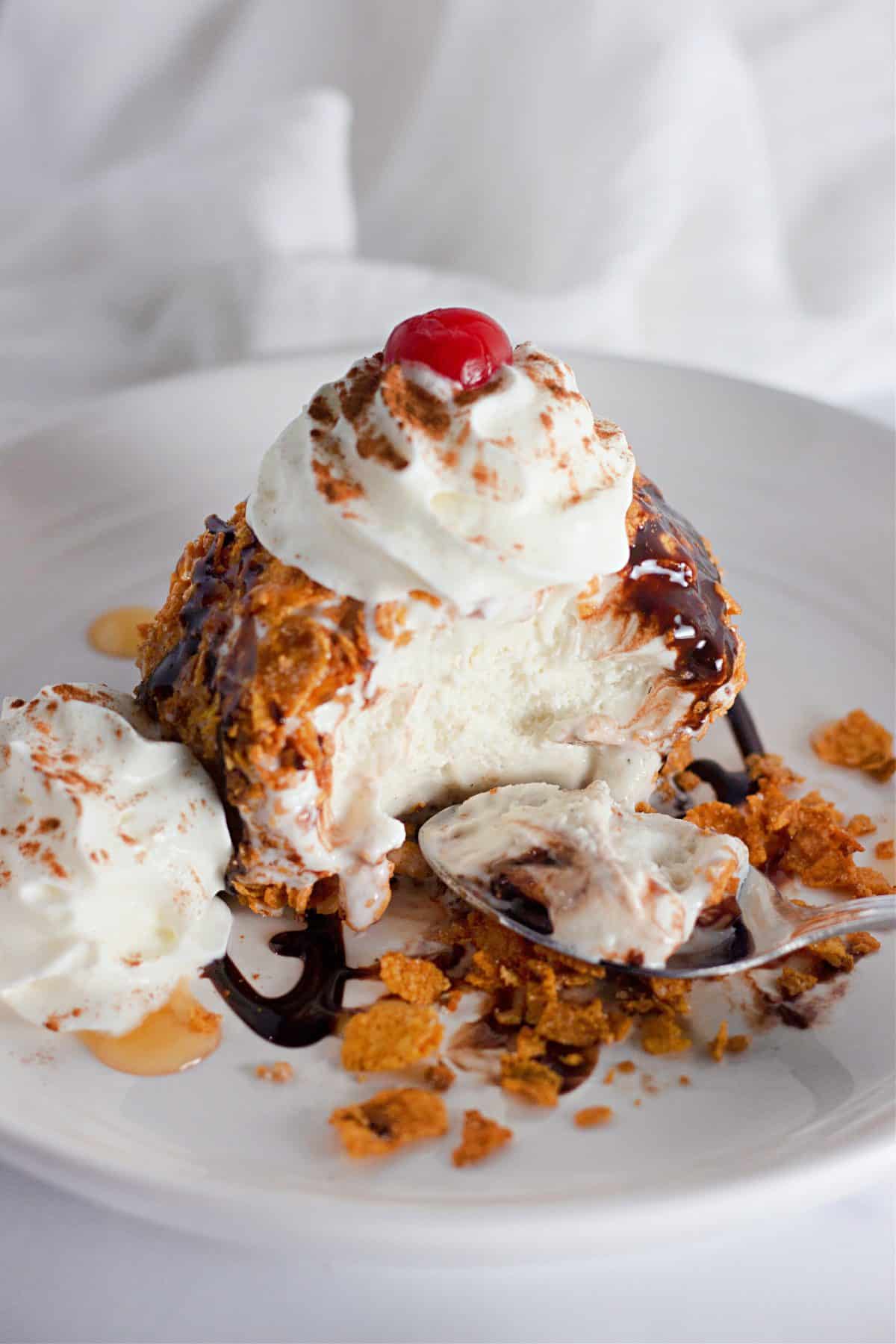 fried ice cream topped with a drizzle of chocolate sauce, whipped cream, and a a maraschino cherry on a small white plate