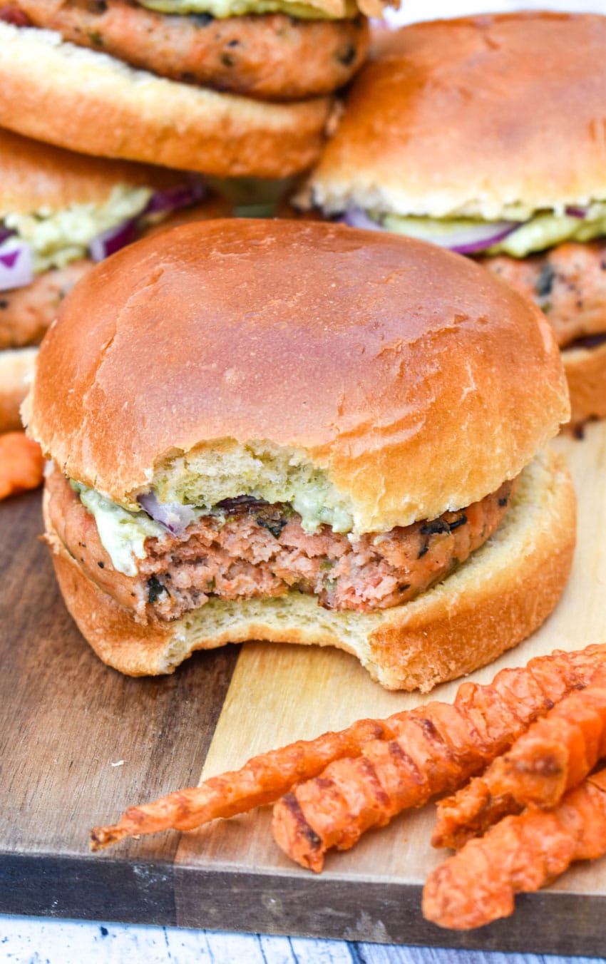 a bitten grilled salmon burger with spinach on a wooden cutting board with sweet potato fries on the side