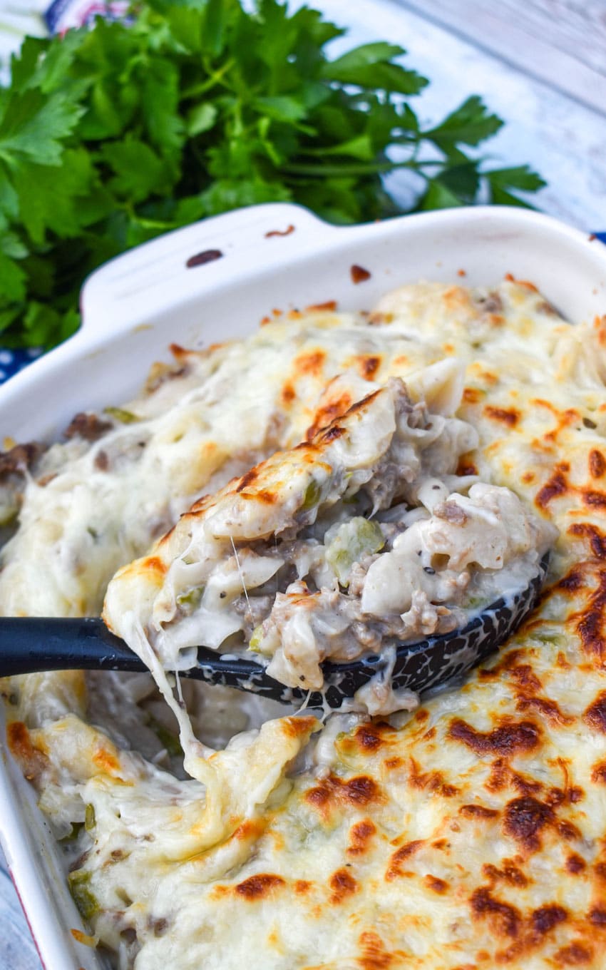 a black spoon scooping philly cheesesteak mac and cheese out of a white casserole dish