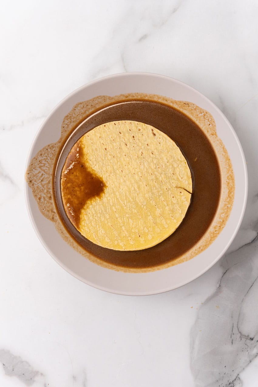 a corn tortilla sitting in birria sauce on a white plate