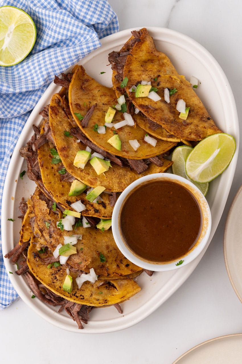 instant pot birria tacos topped with avocado pieces and diced white onion arranged in a row on a white serving platter