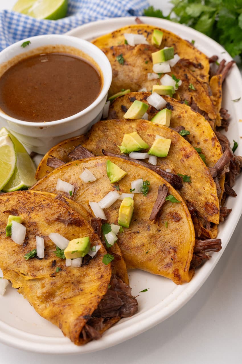 instant pot birria tacos topped with avocado pieces and diced white onion arranged in a row on a white serving platter